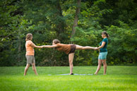 photo of 3 women in yoga shorts, 2 are helping the 3rd do a warrior pose